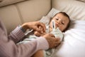 Worried young mother sitting on sofa beside her sick son with high fever. Mom measures temperature using thermometer of Royalty Free Stock Photo