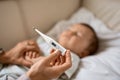 Worried young mother sitting on sofa beside her sick son with high fever. Mom measures temperature using thermometer of Royalty Free Stock Photo