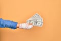 Wounded hand of caucasian young woman cover with bandage holding bunch of dollars banknotes over isolated yellow background