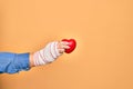 Wounded hand of caucasian young woman cover with bandage holding red heart over isolated yellow background