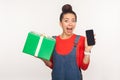 Wow, gift for mobile user! Portrait of amazed joyful pretty girl with hair bun in denim overalls holding present box and cell Royalty Free Stock Photo