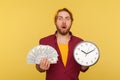 Wow, time to make money! Amazed hipster bearded guy in checkered shirt holding big clock and dollars banknotes