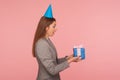 Wow, unexpected present! Side view of surprised woman in business suit and with party cone on her head holding gift box