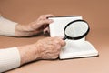 Wrinkled hands of a senior woman are reading a book with magnifier glass. Beige background. The concept of investigation