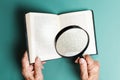 Wrinkled hands of a senior woman are reading a book with magnifier glass. Flat lay. Turquoise background. The concept of