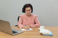 Writing After Retirement. Asian senior woman writer is writing details on book while working on laptop at home Royalty Free Stock Photo