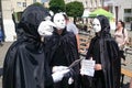 Three actors, dressed in white theater masks and black cloaks, entertain people at a festive fair on a city street.