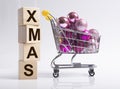 Xmas inscription on cubes near shopping cart or trolley with pink christmas ornaments on white background, side view Royalty Free Stock Photo