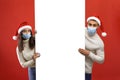 Xmas offer. Young arab couple in Santa hats and medical face masks holding white advertisement board, red background