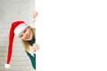 Xmas Santa blonde woman peeking from behind blank sign billboard assign white brick wall. Christmas gnome girl, copy Royalty Free Stock Photo