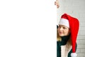 Xmas Santa blonde woman peeking from behind blank sign billboard assign white brick wall. Christmas gnome girl, copy Royalty Free Stock Photo