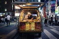 Yakitori stall: woman cooking and selling skewers in a van at night. People walking on the street, eating street food. Shinjuku,