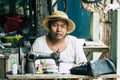 Yangon, Myanmar - March 2019: Asian man in straw hat behind mechanical sewing machine in the street tailor shop