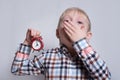 Yawning little blond boy with a red alarm clock in his hands. Morning concept. Light background