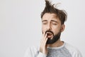 Yawning and sleepy bearded guy with messy hair covering mouth with hand, standing over gray background. Man overslept Royalty Free Stock Photo