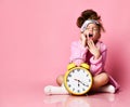 Yawning teenage girl holding an alarm clock while sitting in a housecoat on the floor.