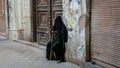 Old Iranian muslim woman wearing chador, the black dress, covering her face, sitting by a very old building