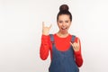Yeah, that`s crazy! Portrait of delighted stylish pretty girl with hair bun in denim overalls showing rock and roll gesture