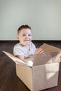 A 4 year old blonde boy sits in a box at home and looks at the camera.