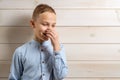 A 10-year-old boy in a blue shirt smiles on a light wooden background and covers his nose with his hands because of an unpleasant