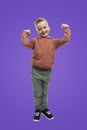 A 5-year-old boy stands and shows muscles in his arms. A child in jeans and a brown sweater. Sports and children`s health. Purple