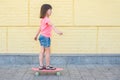 A 7-8-year old girl in a pink T-shirt is skateboarding at the yellow wall background. Lifestyle Royalty Free Stock Photo