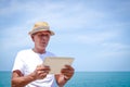 An elderly man comes to rest at the sea, holding a tablet to view information on the internet.