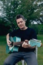 40-50 year old man with a tupe and gray hair playing an acoustic guitar in a garden.