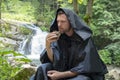 A 35-40-year-old monk in a black cassock with a hood eats bread against the background of a mountain landscape with a waterfall. C