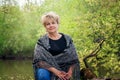 55-60-year-old woman in a knitted shawl is sitting on the riverbank. woman is resting in nature alone Royalty Free Stock Photo