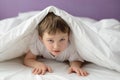 7 years old boy hiding in bed under a white blanket or coverlet Royalty Free Stock Photo