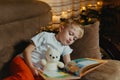 3-4 years old boy is reading a book sitting on a sofa with teddy bear. Home reading of fairy tales. Cute little boy is Royalty Free Stock Photo