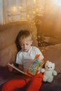 3-4 years old boy is reading a book sitting on a sofa with teddy bear. Home reading of fairy tales. Cute little boy is Royalty Free Stock Photo