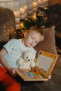 3-4 years old boy is reading a book sitting on a sofa with teddy bear. Home reading of fairy tales. Cute little boy is Royalty Free Stock Photo