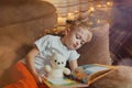 3-4 years old boy is reading a book sitting on a sofa with teddy bear. Home reading of fairy tales. Cute little boy is Royalty Free Stock Photo