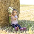 3 years old little girl with a bouquet of daisies sits by a sheaf of straw on a sunny