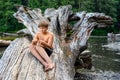 Young boy sitting on a fallen tree trunk, and focusing on to carving a wooden stick for the grill with a small knife