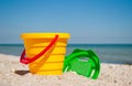 Yellow baby bucket with a red handle and a plastic green sieve left on a blue sea background sand summer sunny day, baby toys Royalty Free Stock Photo