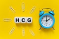 On a yellow background, a blue alarm clock, paper clips and white cubes on which the text is written - HCG