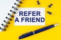 On a yellow background lies a notebook, a pen and a business card with the inscription - Refer a friend