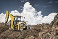 A yellow backhoe loader works at a construction site, digging into the ground while fluffy clouds loom in the bright afternoon sky Royalty Free Stock Photo