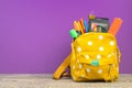 Yellow backpack with white polka dots with different colorful stationery on table. Purple background. Back to school