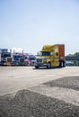 Yellow big rig semi truck with orange semi trailer driving on the truck stop driving out of a line of parked semi trucks Royalty Free Stock Photo