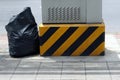 A yellow and black cement base warning sign of a telephone exchange booth with garbage bag on the sidewalk