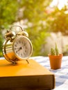 Yellow book with vintage alarm clock and cactus on blue plaid tablecloth. The background is green from tree and light bokeh Royalty Free Stock Photo