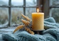 A yellow candle atop a wooden holder, wrapped in a soft blue knit blanket with delicate dried flowers by a frosted window