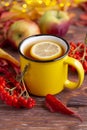 Yellow cup of tea with slice of lemon, colorful red and yellow autumn leaves, an apple and bunch of rowanberry.