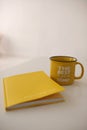 A yellow diary with a yellow mug stands on the edge of a white table in a white room.