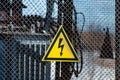 Yellow electricity warning sign with lightning hanging on gray wire mesh fence that surrounds transformer
