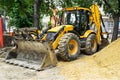 Yellow excavator with a bucket and large heap of sand at a road construction site on a city street on a summer day.